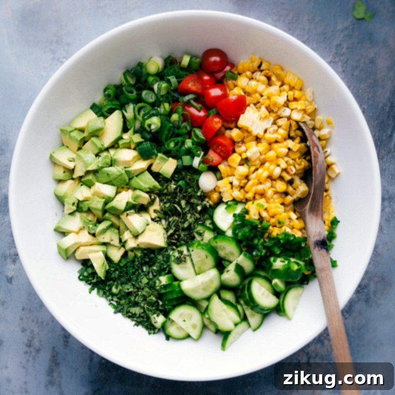 A clear glass bowl filled with all the fresh, prepped ingredients for the corn salad recipe. This includes bright yellow corn kernels, diced red cherry tomatoes, thinly sliced green onions, chopped green cucumbers, and fresh herbs like cilantro and basil, all ready for mixing and dressing.
