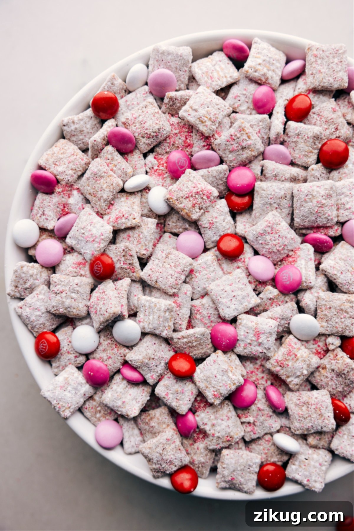 Our Favorite Valentine Muddy Buddies, Perfectly Coated and Ready to Enjoy A close-up shot of Valentine Muddy Buddies in a bowl, showcasing the generous coating of powdered sugar, along with various red, pink, and white Valentine's M&Ms scattered throughout.