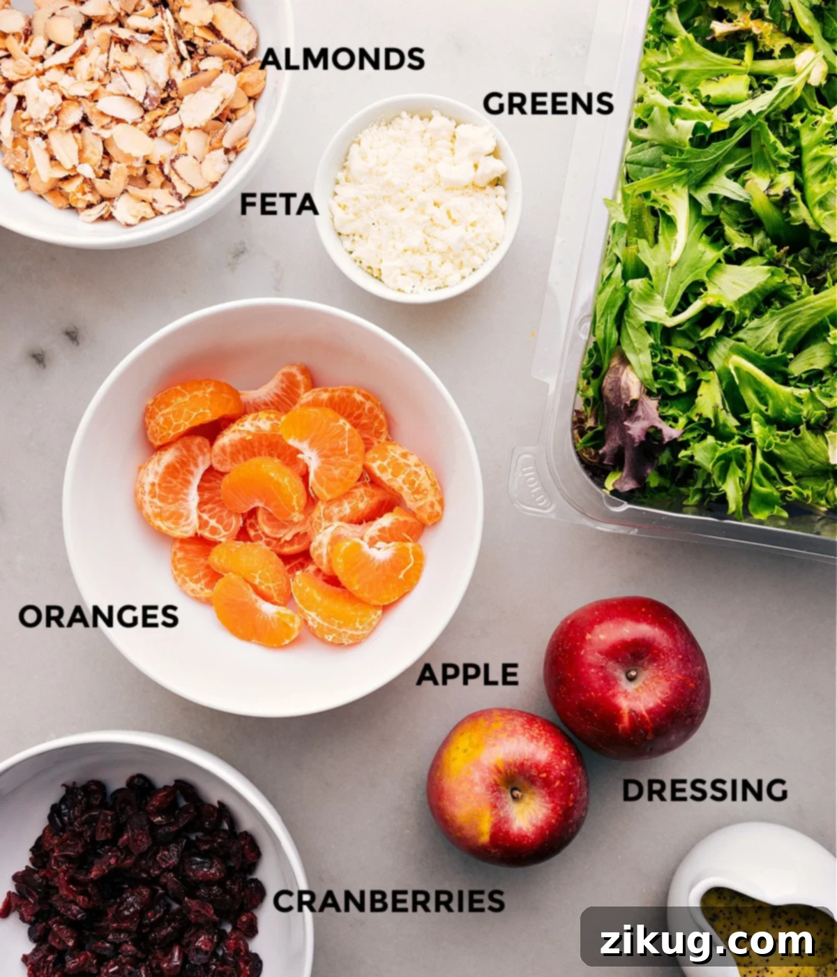 An array of fresh, colorful ingredients laid out, including crisp greens, segmented oranges, sliced apples, dried cranberries, feta cheese, and homemade candied almonds, ready for the mandarin orange salad assembly.
