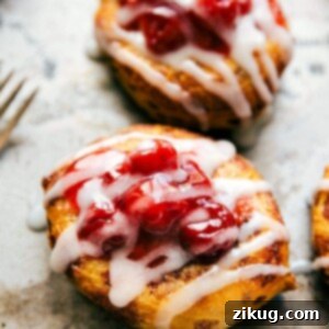 A close-up of a single Cinnamon Roll Cherry Danish, perfectly baked with its cream cheese and cherry filling, drizzled with white icing.