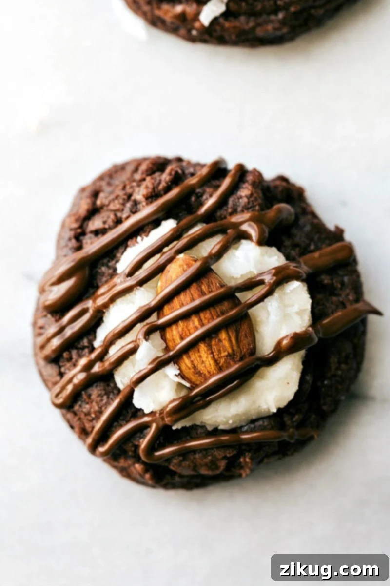 Delicious Almond Joy Cookies featuring a rich brownie base, creamy coconut frosting, roasted almonds, and a chocolate drizzle. A perfect dessert, quick and easy to make. #dessert #best #quick #easy #simple #chocolate #frosting #almondjoy #thumbprint #cookies Up close overhead image of an Almond Joy Cookie showing rich chocolate, white coconut, and a whole almond
