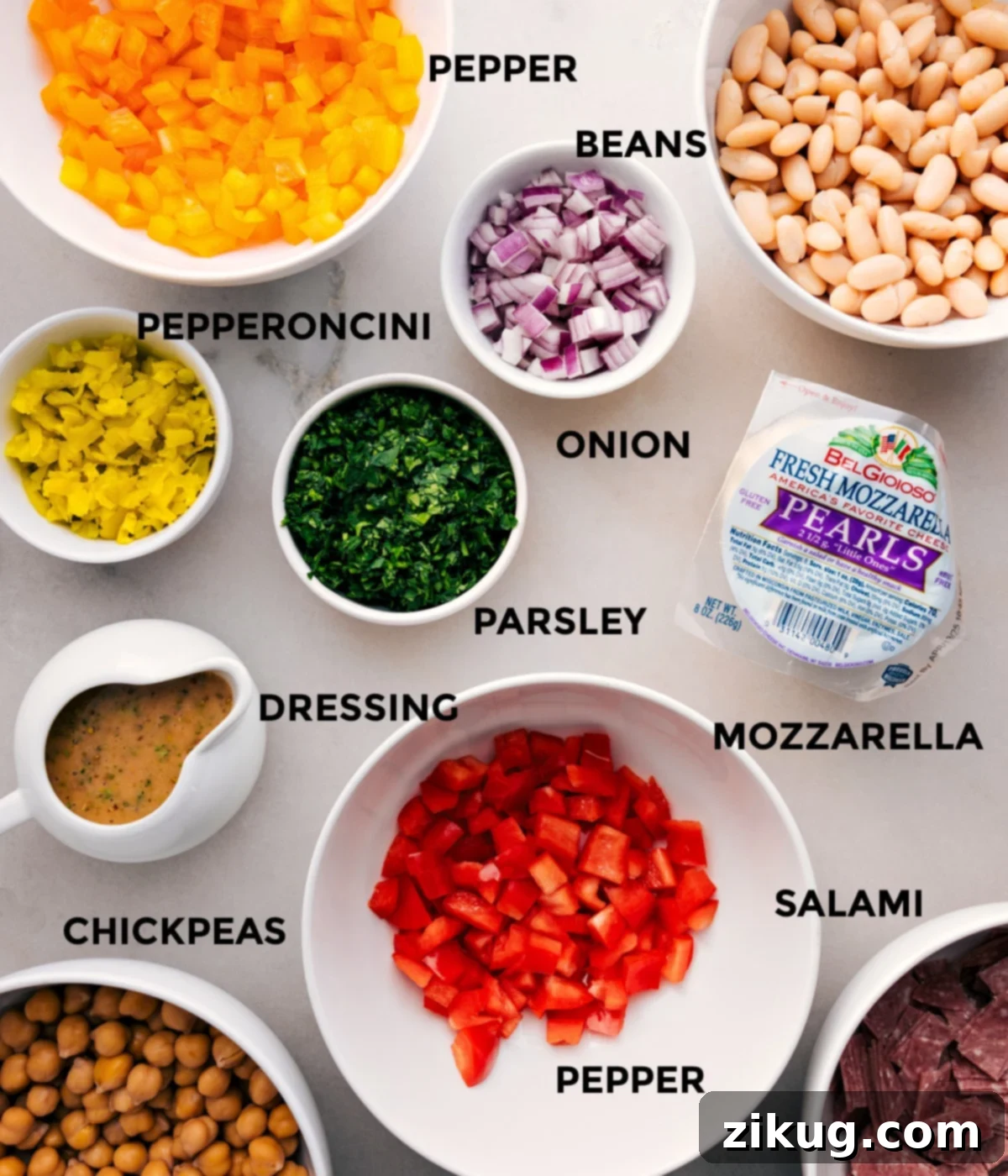 All the vibrant ingredients for a delicious dense bean salad are prepped and neatly arranged for easy assembly: a mix of beans, chopped onion, fresh parsley, mozzarella pearls, spicy pepper salami, tangy pepperoncini, and a jar of homemade dressing.