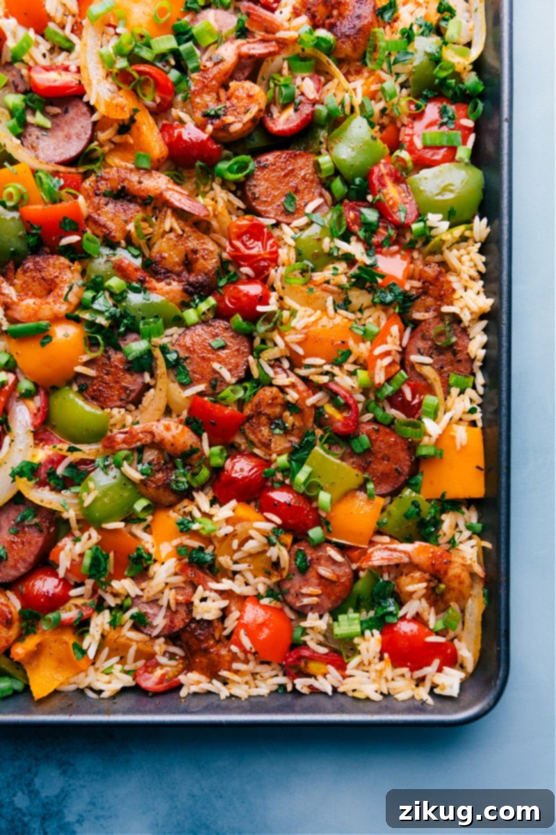 One-Pan Jambalaya is an easy, nutritious, 30-minute meal of oven-roasted & seasoned sausage, shrimp, veggies, and rice. Everything gets cooked together on one sheet pan! Recipe via ChelseasMessyApron #dinner #quick #easy #simple #best #veggies #shrimp #sausage #rice #onepan #jambalaya Overhead view of a finished One-Pan Jambalaya, showcasing colorful bell peppers, sausage, shrimp, and rice with fresh green onions and parsley.