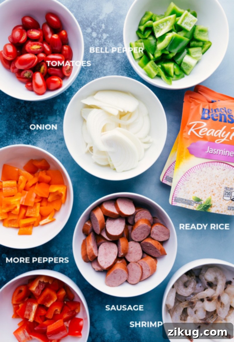 One-Pan Jambalaya is an easy, nutritious, 30-minute meal of oven-roasted & seasoned sausage, shrimp, veggies, and rice. Recipe via ChelseasMessyApron #dinner #quick #easy #simple #best #veggies #shrimp #sausage #rice #onepan #jambalaya A collection of fresh ingredients for One-Pan Jambalaya, including bell peppers, onions, sausage, shrimp, cherry tomatoes, rice packets, and various spices.