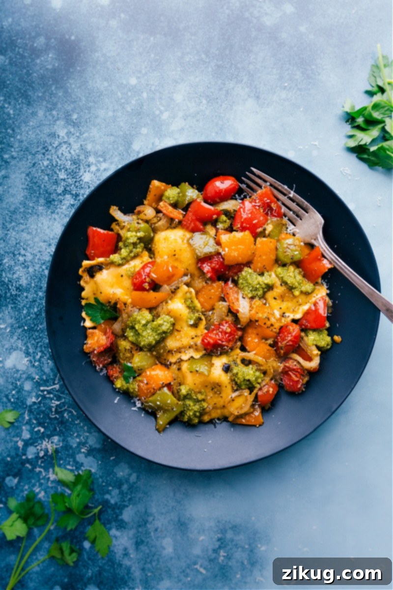 Pesto Ravioli combines perfectly caramelized roasted veggies with tender cheese-filled ravioli. Top each bowl with a drizzle of olive oil, lots of freshly grated Parmesan cheese, and another spoonful of pesto for an unforgettable pasta dish! Recipe via ChelseasMessyApron #dinner #quick #easy #simple #roasted #veggies #onetray #tomatoes #pesto #ravioli A bowl of Pesto Ravioli with roasted vegetables, garnished with fresh basil leaves and grated Parmesan cheese.