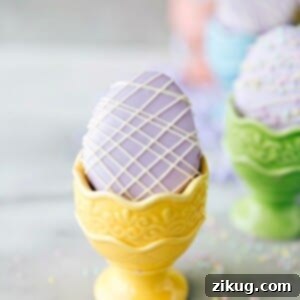 Delicate mini egg-shaped cakes, frosted in pastel colors and intricately decorated with white chocolate drizzle and festive Easter sprinkles.