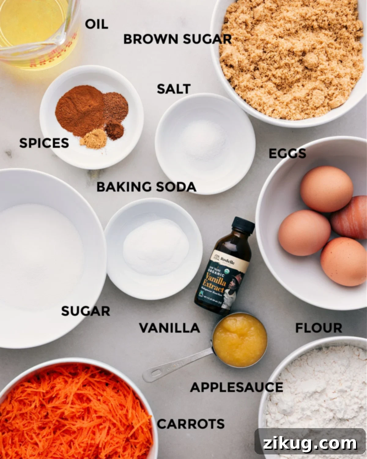 A beautifully arranged collection of fresh, high-quality ingredients laid out for making the ultimate carrot cake: including vegetable oil, brown sugar, eggs, essential salt, a vibrant selection of warm spices, baking soda, pure vanilla extract, granulated sugar, all-purpose flour, unsweetened applesauce, and freshly shredded carrots.