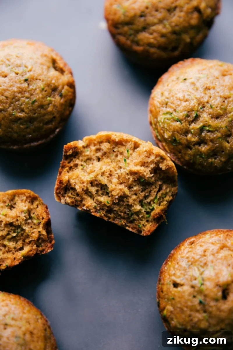 A close-up view of two freshly baked healthy zucchini muffins, one whole and one split in half to showcase its tender, moist interior dotted with zucchini flecks. They sit on a wooden surface.