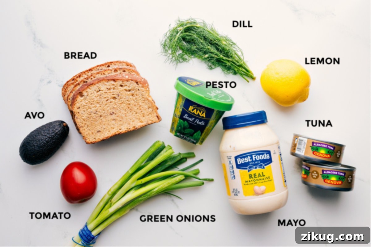 All the ingredients prepped for easy assembly of the Tunacado sandwich, including avocado slices, fresh tomato, thin bread, chopped green onions, vibrant pesto, fresh dill sprigs, a lemon, canned tuna, and mayonnaise.