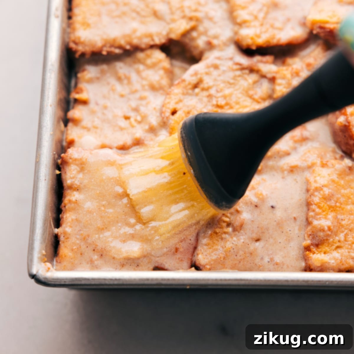 Cozy Classic Bread Pudding 5 A pastry brush glides across the top of the bread pudding in a baking pan, evenly coating it with melted butter to achieve a golden, crispy crust during baking.
