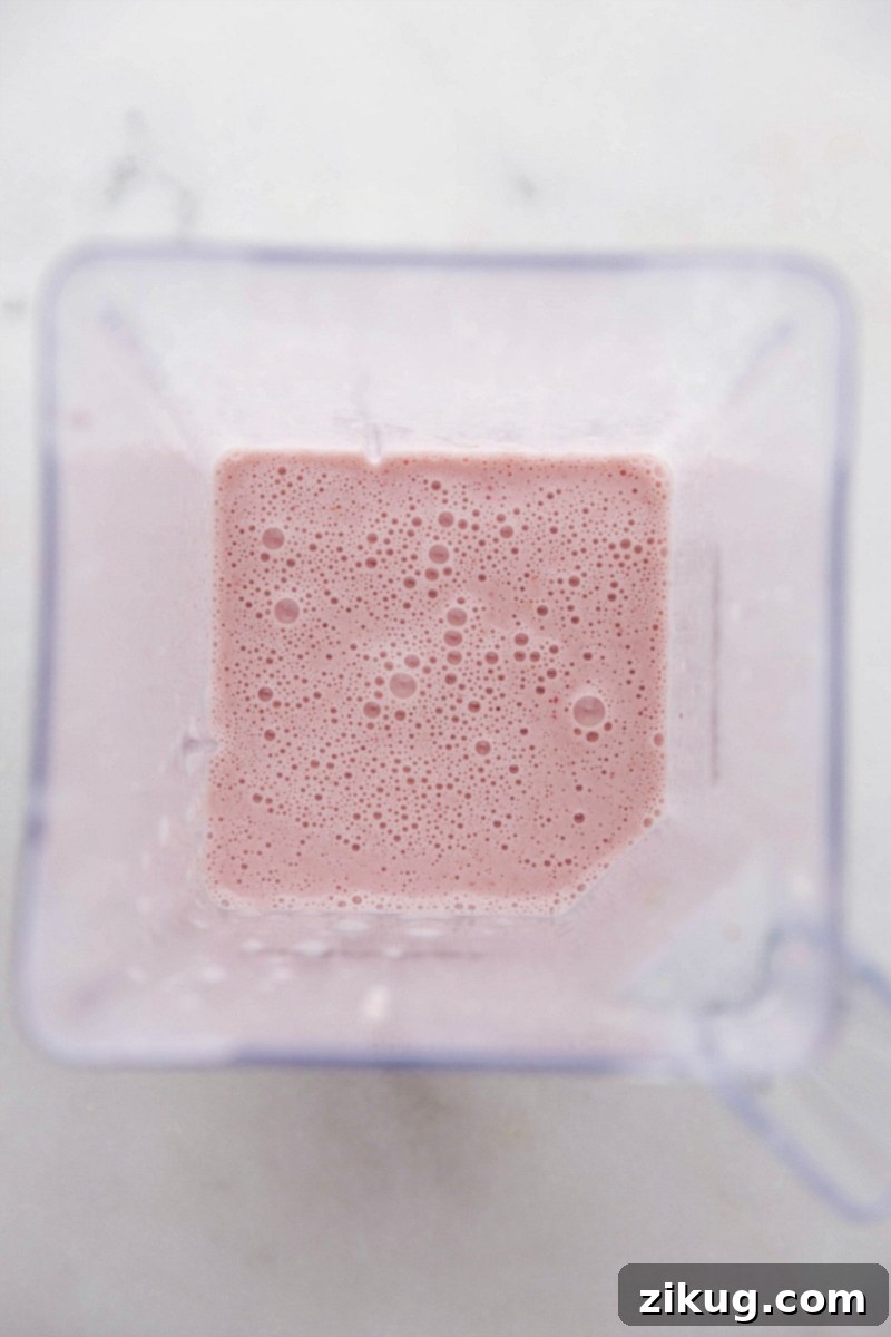 The transformation is complete! All five essential ingredients – frozen strawberries, frozen banana, milk, Greek yogurt, and honey – are blended to creamy perfection in this delicious, healthy, and easy Strawberry Banana Smoothie. Ready to be poured and enjoyed immediately. Overhead process shot of the ingredients for Strawberry Banana Smoothies, perfectly blended into a smooth, creamy texture within a high-powered blender.