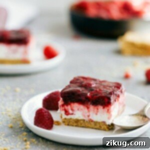 A top-down view of a rectangular Raspberry Jello Dessert bar, showing its distinct layers: a brown graham cracker crust, a creamy white cheesecake filling, and a vibrant red raspberry Jello topping.