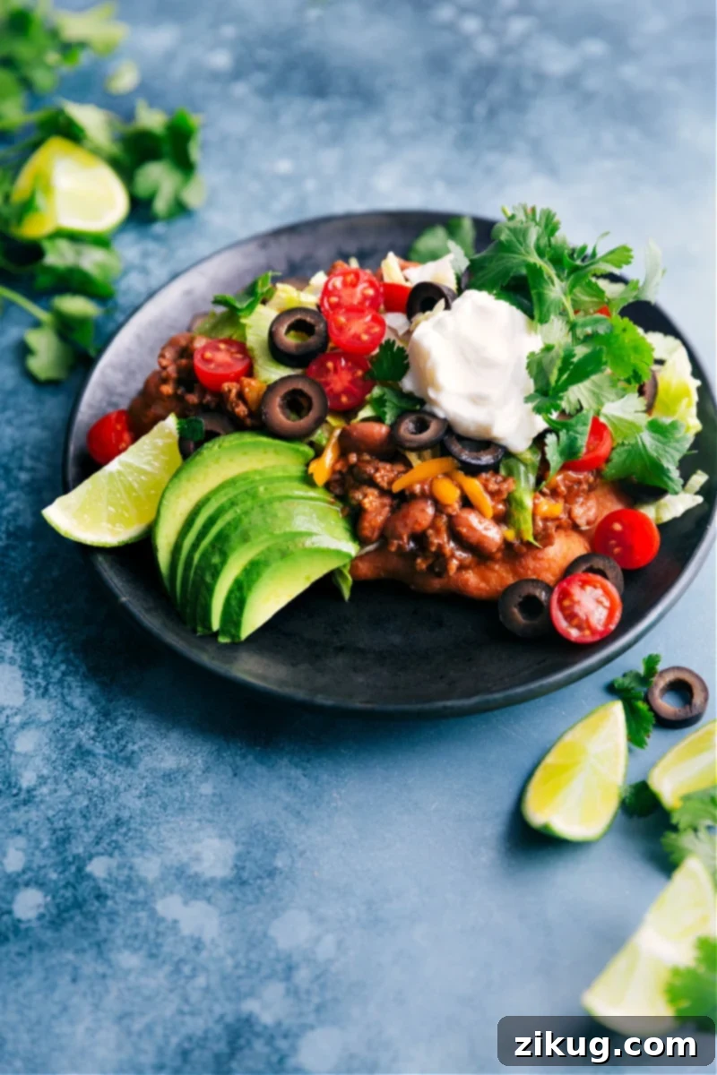 These Navajo Tacos start with the absolute best homemade Fry Bread! The bread is topped with a deliciously seasoned beef and bean mixture and then topped with all your favorite taco toppings like cheese, lettuce, tomatoes, and sour cream. Recipe via ChelseasMessyApron #dinner #quick #easy #simple #best #avocado #toppings #familyfriendly #kidfriendly #frybread #navajo #tacos A delicious Navajo Taco presented on a plate, showcasing its rich toppings on golden Fry Bread.