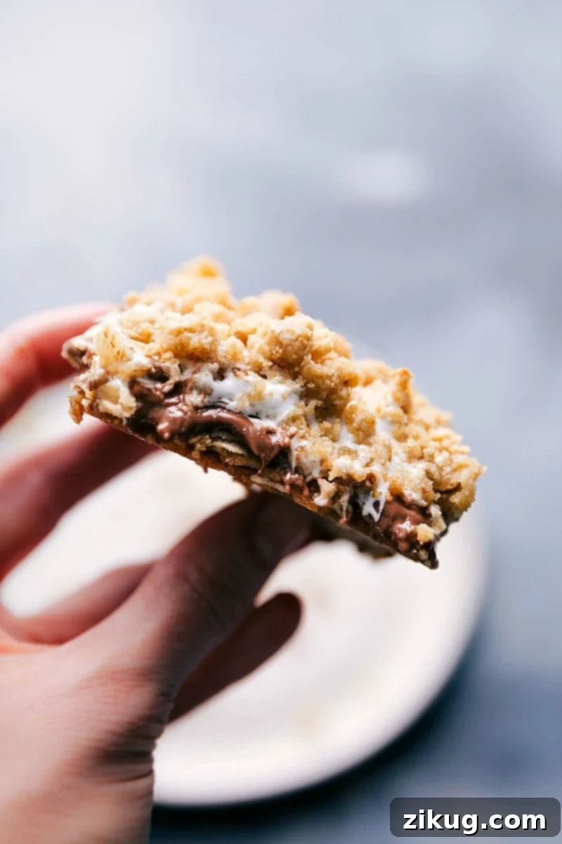 A hand holds a freshly baked S'mores Cookie Bar, showcasing its incredibly gooey, melted marshmallow, and chocolate-filled center, tempting viewers with its irresistible texture.