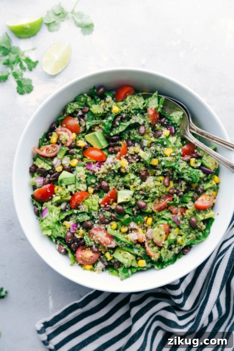 A large, colorful bowl overflowing with a healthy Southwest Quinoa Salad, featuring a generous mix of quinoa, black beans, corn, tomatoes, avocado, and green leafy vegetables. Two serving spoons rest on the side, inviting you to dig in.