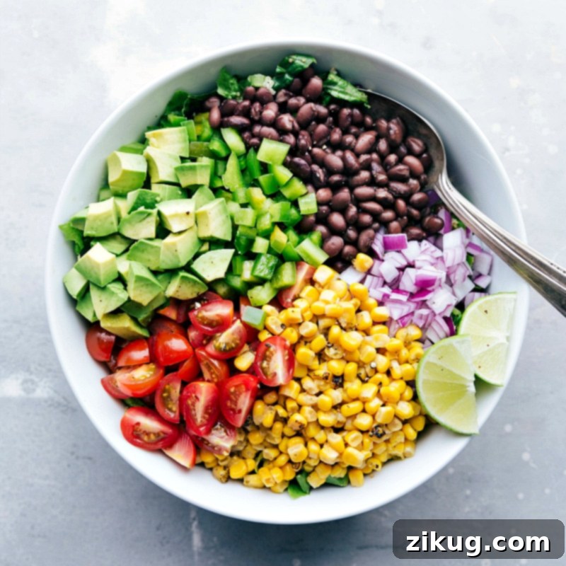 A close-up shot of a large mixing bowl where remaining ingredients like black beans, vibrant roasted corn, chopped red onions, cherry tomatoes, and diced green bell peppers are being added to a base of cooked quinoa and crisp romaine lettuce, illustrating the assembly process of the salad.