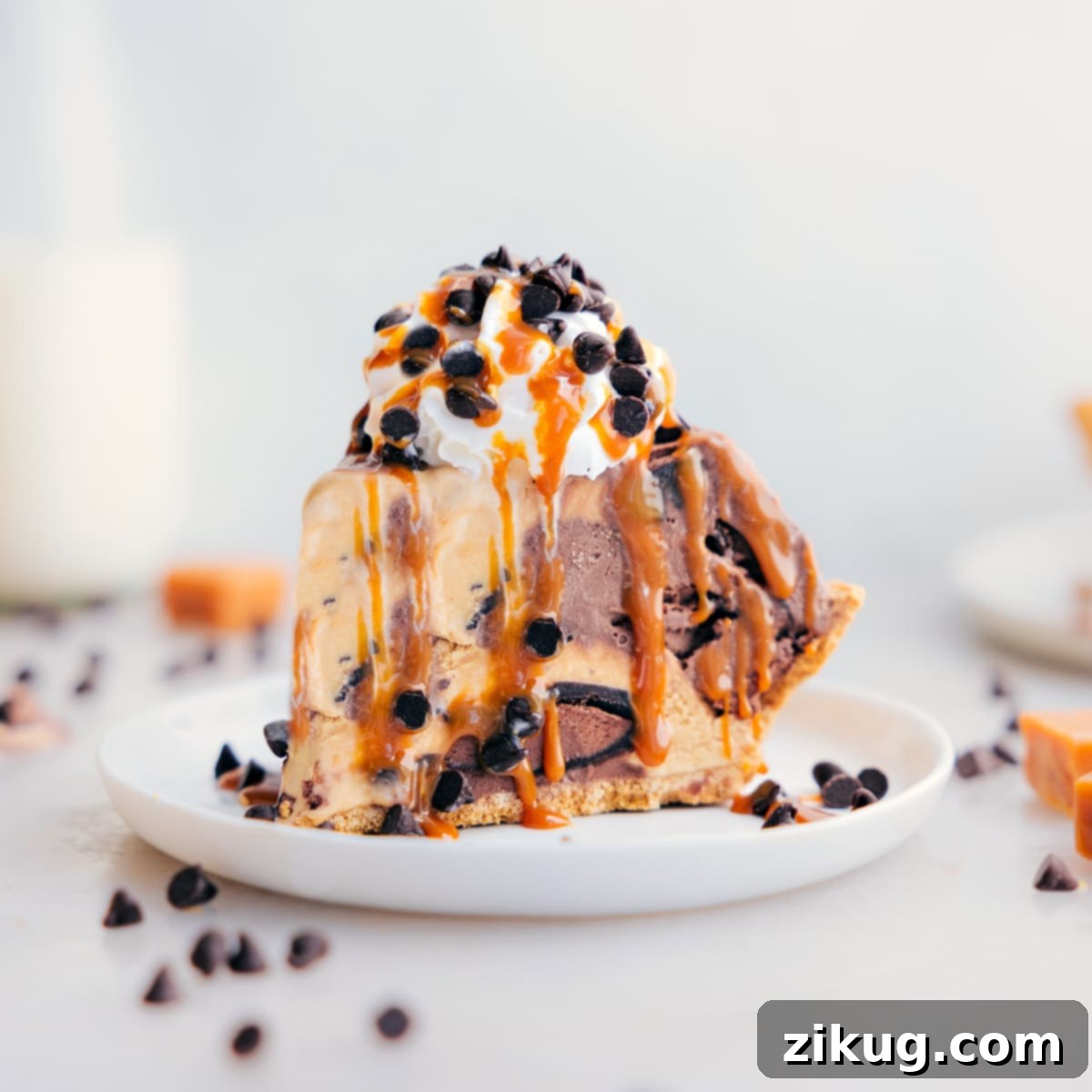 Decadent And Delicious Ice Cream Pie Recipe A delectable slice of ice cream pie on a white plate, showcasing the beautifully layered chocolate and caramel ice cream, topped with whipped cream, a caramel drizzle, and chocolate chips, against a blurred background of the full pie.