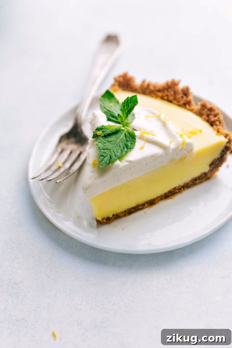 A pristine slice of creamy lemon pie, showcasing its golden graham cracker crust, vibrant yellow filling, and a delicate swirl of whipped cream, garnished with fresh mint on a white plate.