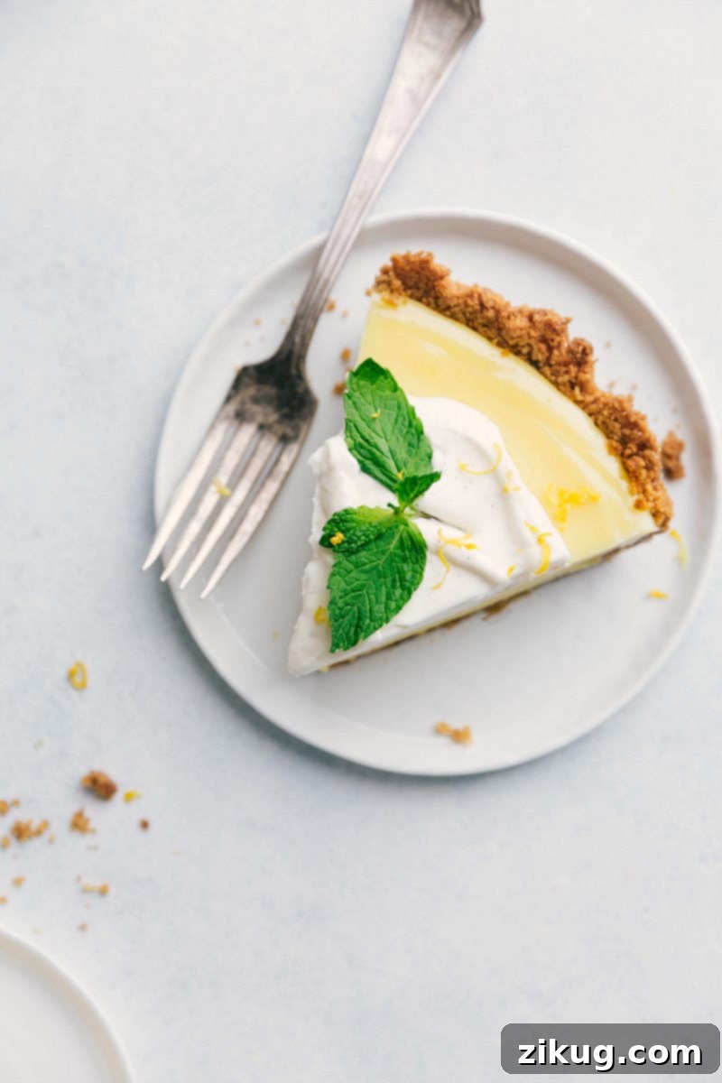 A perfectly portioned slice of vibrant lemon pie on a white plate, highlighting its distinct layers of golden crust, creamy yellow filling, and fluffy white whipped cream, accented with a fresh mint leaf.