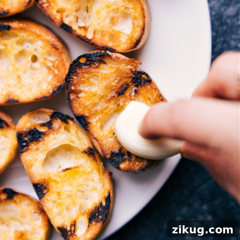 Process shot of a garlic clove being rubbed onto toasted bread slices for bruschetta.