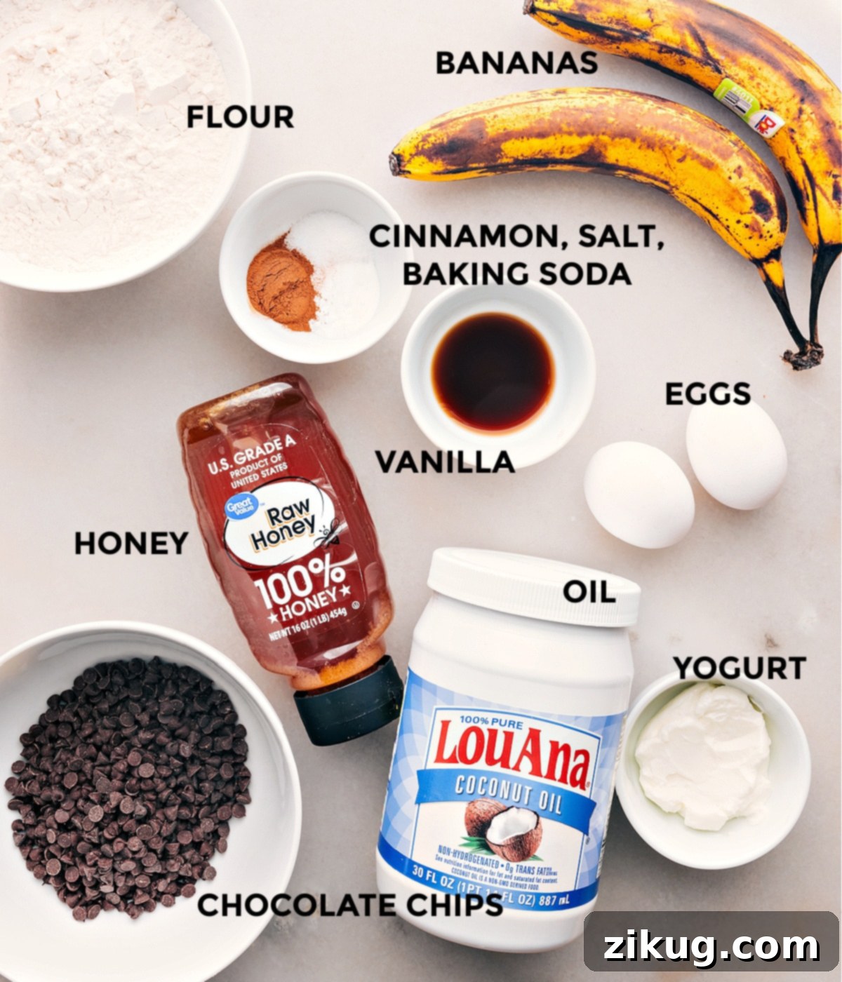 Ingredients In Healthy Banana Bread Recipe A vibrant display of all the healthy ingredients for banana bread: flour, ripe bananas, cinnamon, salt, baking soda, eggs, vanilla extract, honey, chocolate chips, coconut oil, and Greek yogurt, neatly arranged on a counter.