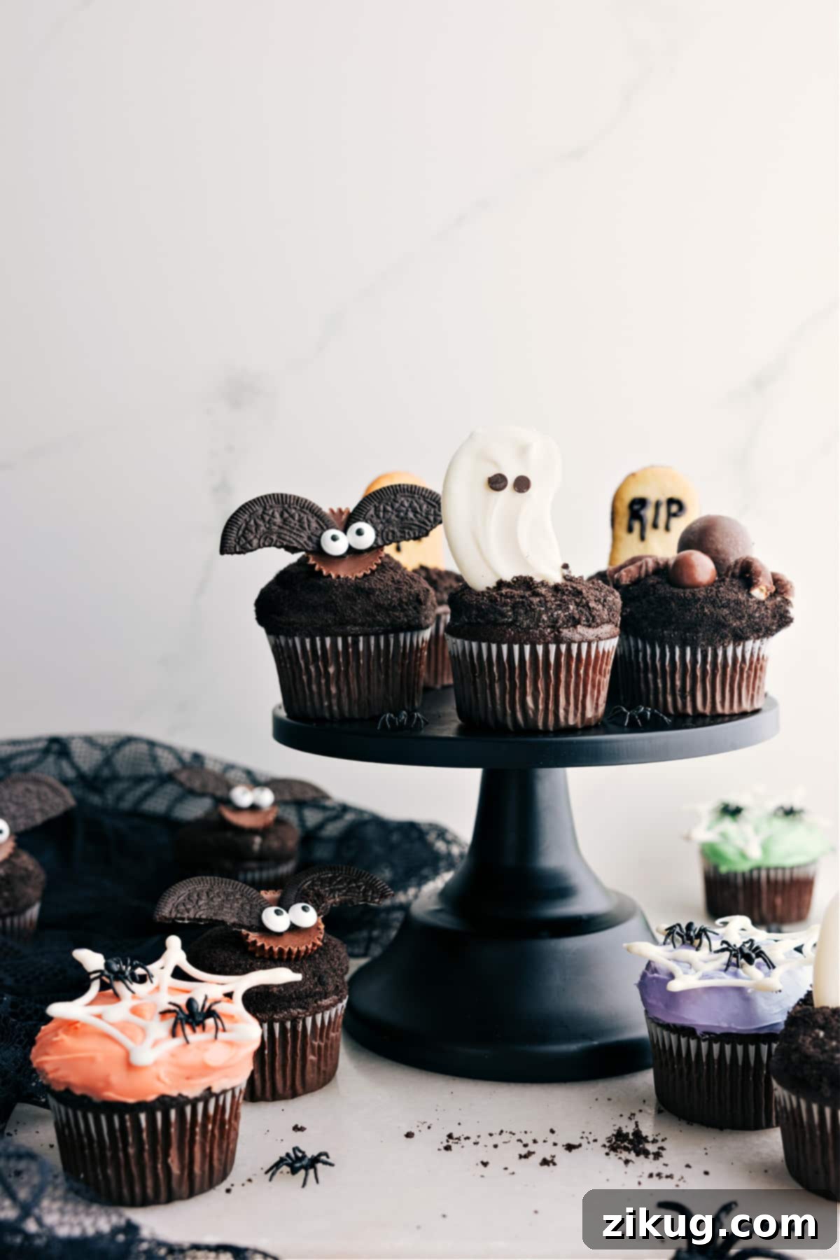 Assorted Halloween cupcakes displayed on a cupcake holder, featuring a ghost, a spider, a bat, and a tombstone cupcake, all against a dark, festive backdrop.
