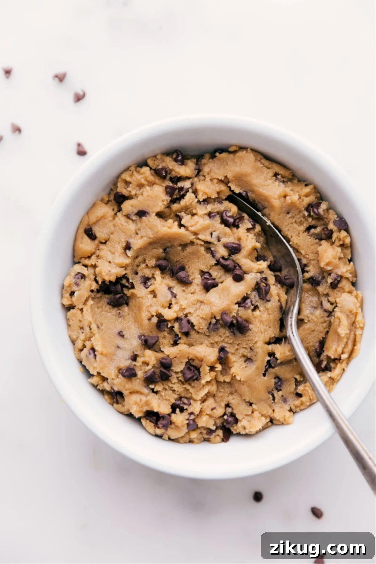 A bowl filled with rich, brown edible chocolate chip cookie dough, generously studded with mini chocolate chips, ready to be enjoyed with a spoon.