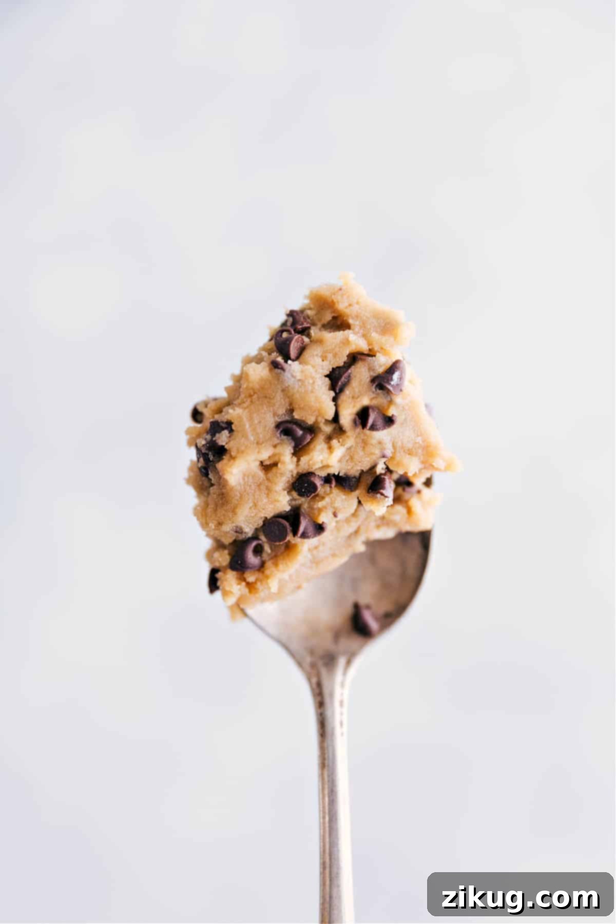 A single spoonful of delicious, rich edible chocolate chip cookie dough is held up against a soft-focus background, showcasing its perfect, creamy texture and abundant mini chocolate chips, inviting a delightful taste.