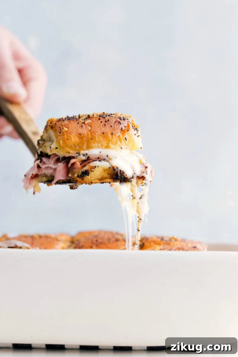 The Best Ham And Cheese Sliders with Cheesy Pull A hand picking up a ham and cheese slider with a satisfying cheesy pull.