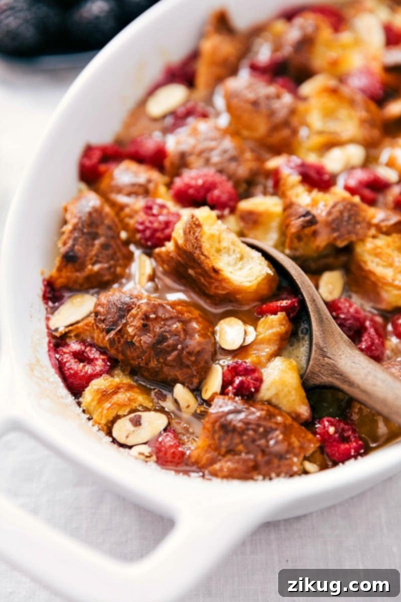 Golden Croissant Bread Pudding with Vanilla Sauce and Fresh Berries Scooping golden-brown croissant bread pudding from a baking dish, showcasing visible chunks of bread, a generous drizzle of vanilla sauce, and vibrant fresh fruit pieces.