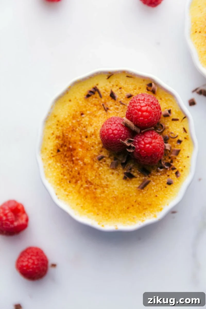 Tuxedo Crème Brûlée is a chocolatey twist on the traditional crème brûlée recipe. This dessert has a chocolate ganache bottom, creamy custard filling, and a caramelized sugar topping. Top these brulée with fresh berries and chocolate curls for a delicious show-stopping dessert. Recipe via ChelseasMessyApron.com #cremebrulee #tuxedo #ganache #French #dessert #elegant Overhead image of the Tuxedo Crème Brûlée garnished with chocolate curls and raspberries.