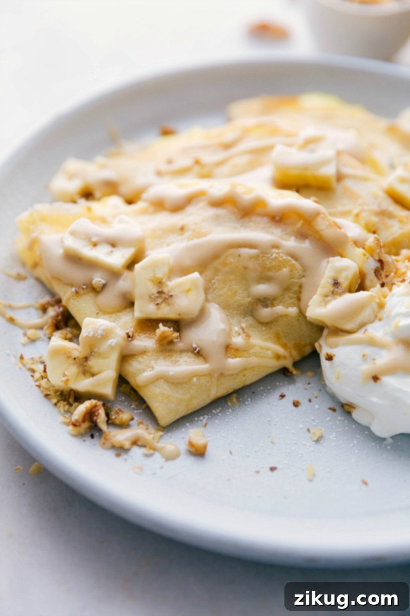 This simple crepe recipe is filled with an easy custard filling, plenty of bananas, and chopped walnuts. Banana Crepes are easy to make -- the actual crepes and custard can be made ahead of time so when you're ready to serve, everything comes together in a flash. Recipe via ChelseasMessyApron.com #crepes #bananas #custart #walnuts #breakfast Overhead image of the ready-to-eat Banana Crepes, beautifully arranged and garnished.