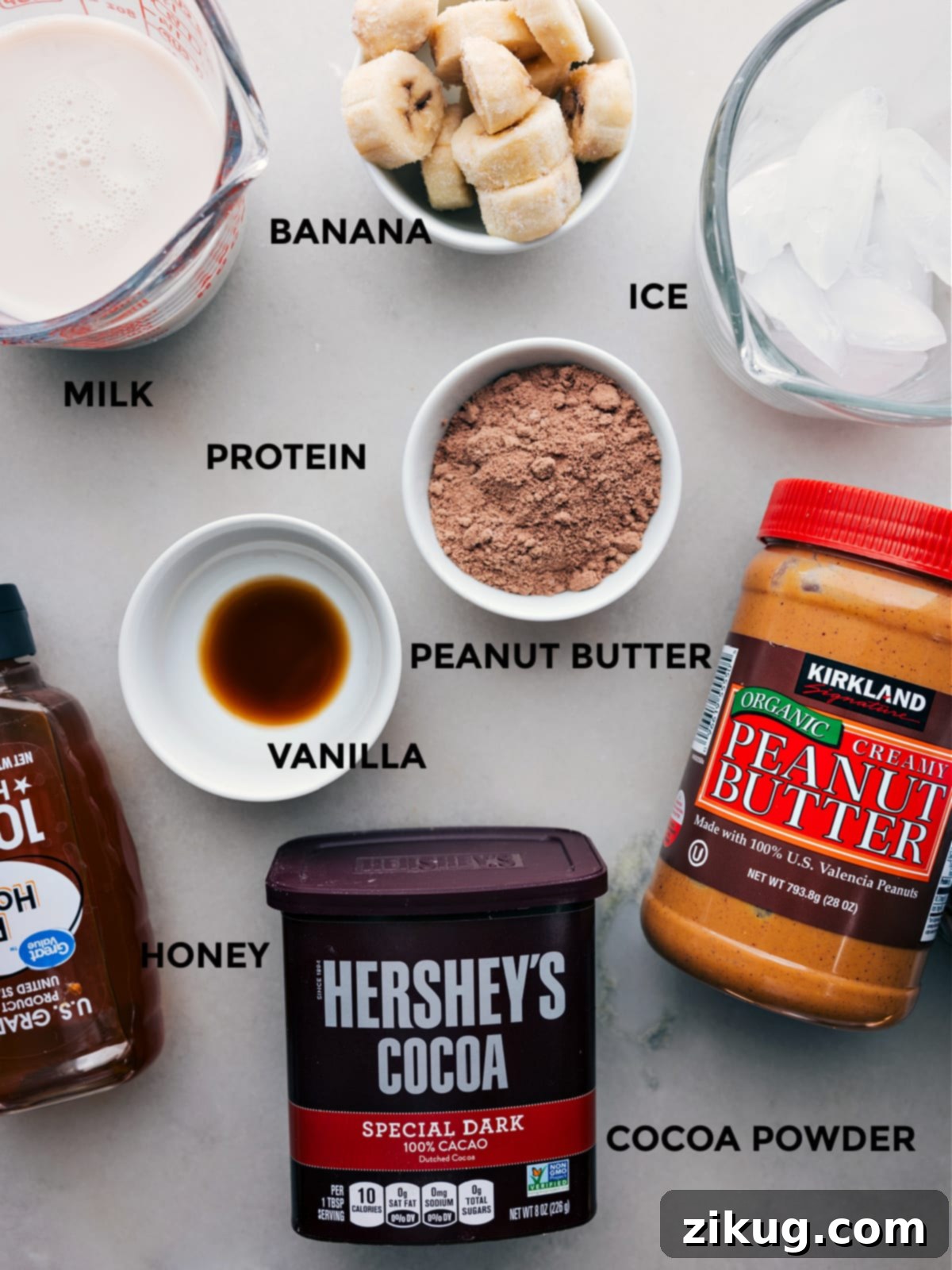 Ingredients In Peanut Butter Protein Shake Recipe All the ingredients including cocoa powder, vanilla, peanut butter, protein powder, bananas, milk, ice, and honey prepped out for easy assembly.