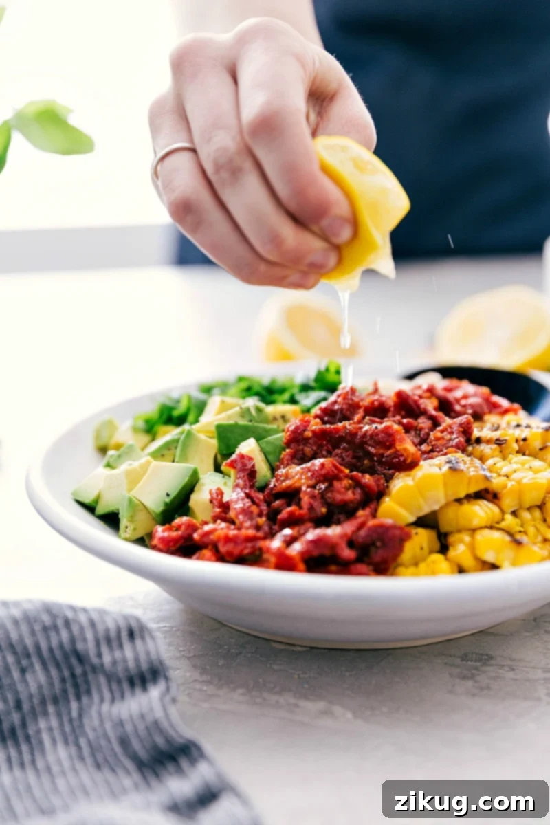 The best ever White Bean Salsa, featuring creamy avocado, crisp grilled corn, savory sun-dried tomatoes, bright fresh lemon, and sweet basil, is our latest culinary obsession! It's absolutely perfect served over grilled Pesto Chicken, mixed into quinoa or couscous for a satisfying vegetarian meal, or enjoyed with crunchy tortilla chips. Recipe from Chelseasmessyapron. #healthy #keto #whole #protein #whole30 #best #avocado #corn #sundriedtomatoes #clean #cleaneating #whitebean #salsa Fresh lemon juice being drizzled over the vibrant White Bean Salsa in a bowl, ready to be served.