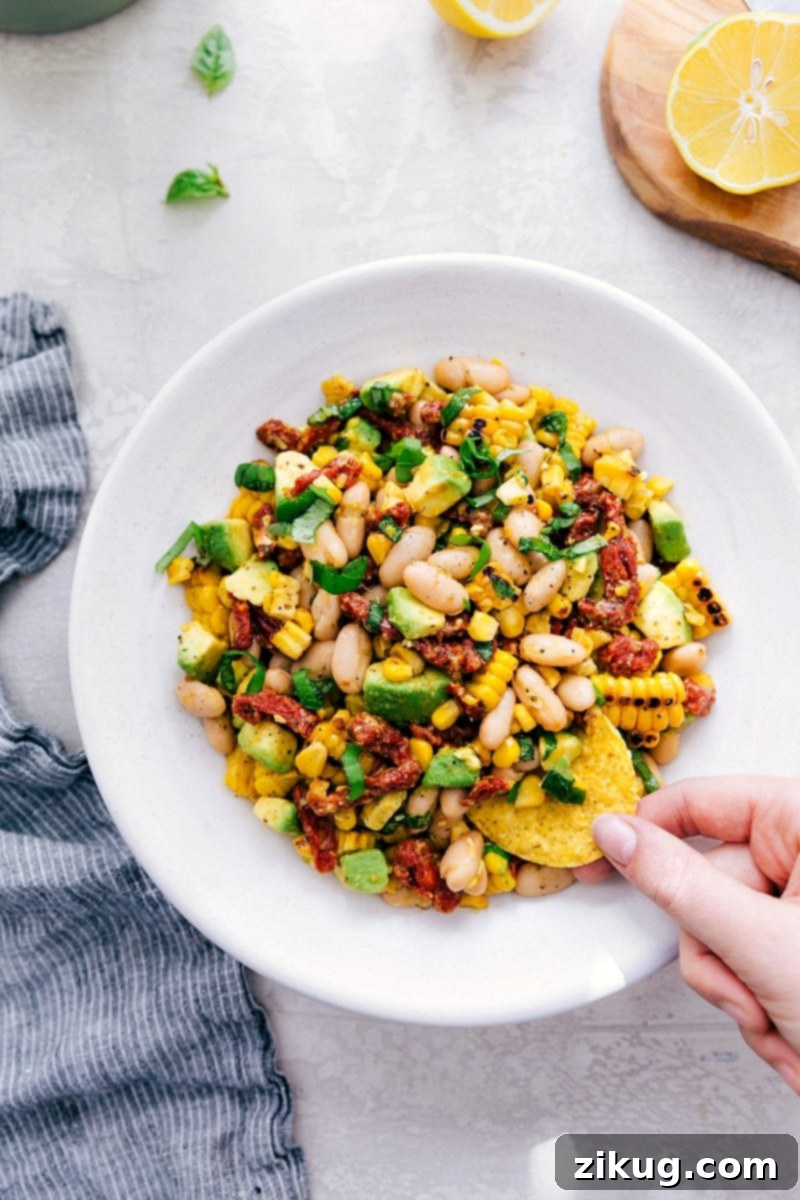 Dive into this delicious White Bean Salsa! Recipe via Chelseasmessyapron. This healthy and satisfying dip is packed with flavor from avocado, corn, sun-dried tomatoes, lemon, and basil. #healthy #keto #whole #protein #whole30 #best #avocado #corn #sundriedtomatoes #clean #cleaneating #whitebean #salsa Overhead shot of White Bean Salsa in a bowl with a hand dipping a chip, emphasizing its appeal.