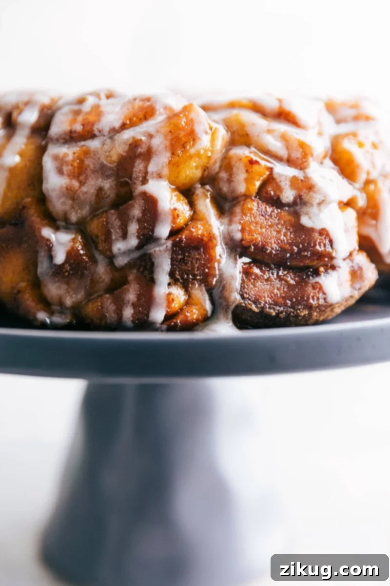 A perfectly baked Cinnamon Roll Monkey Bread, glistening with cream cheese frosting, sits invitingly on a cake stand, ready to be pulled apart and savored. Its golden-brown exterior and visible layers promise a tender, flavorful experience, making it an ideal sweet treat for any occasion.