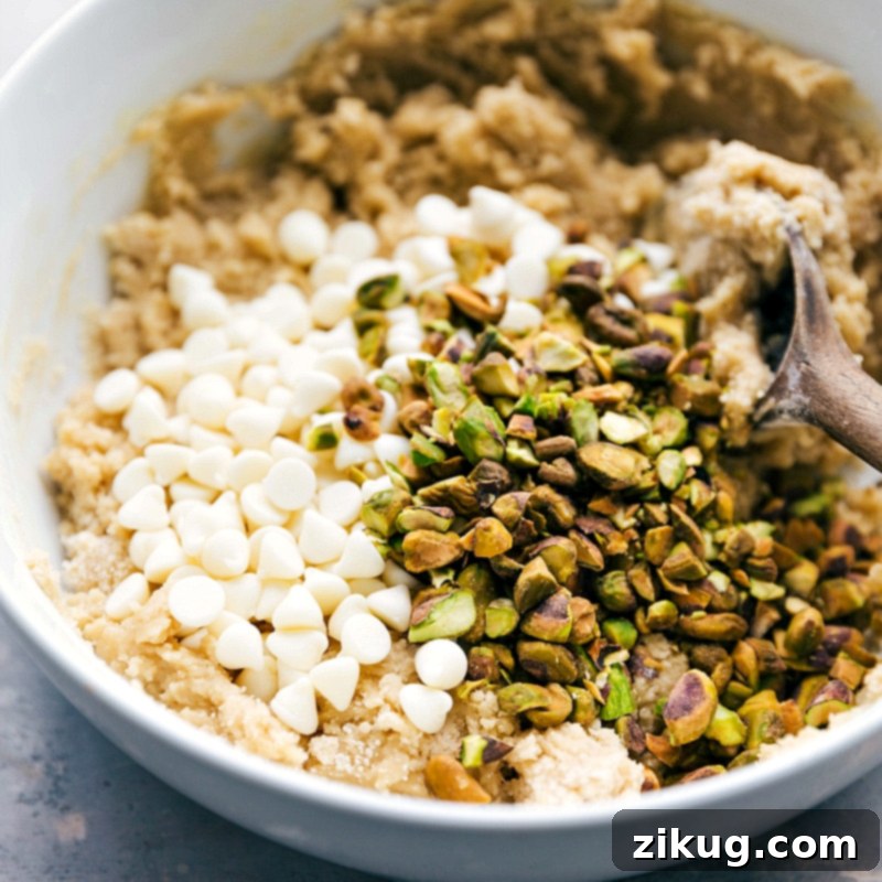 Incorporating White Chocolate Chips and Nuts into Cookie Dough Creamy white chocolate chips and vibrant, coarsely chopped roasted pistachios being added to a bowl of perfectly mixed cookie dough. The ingredients are then gently folded until evenly incorporated, ensuring a delightful blend of flavors and textures in every bite of these homemade pistachio cookies.