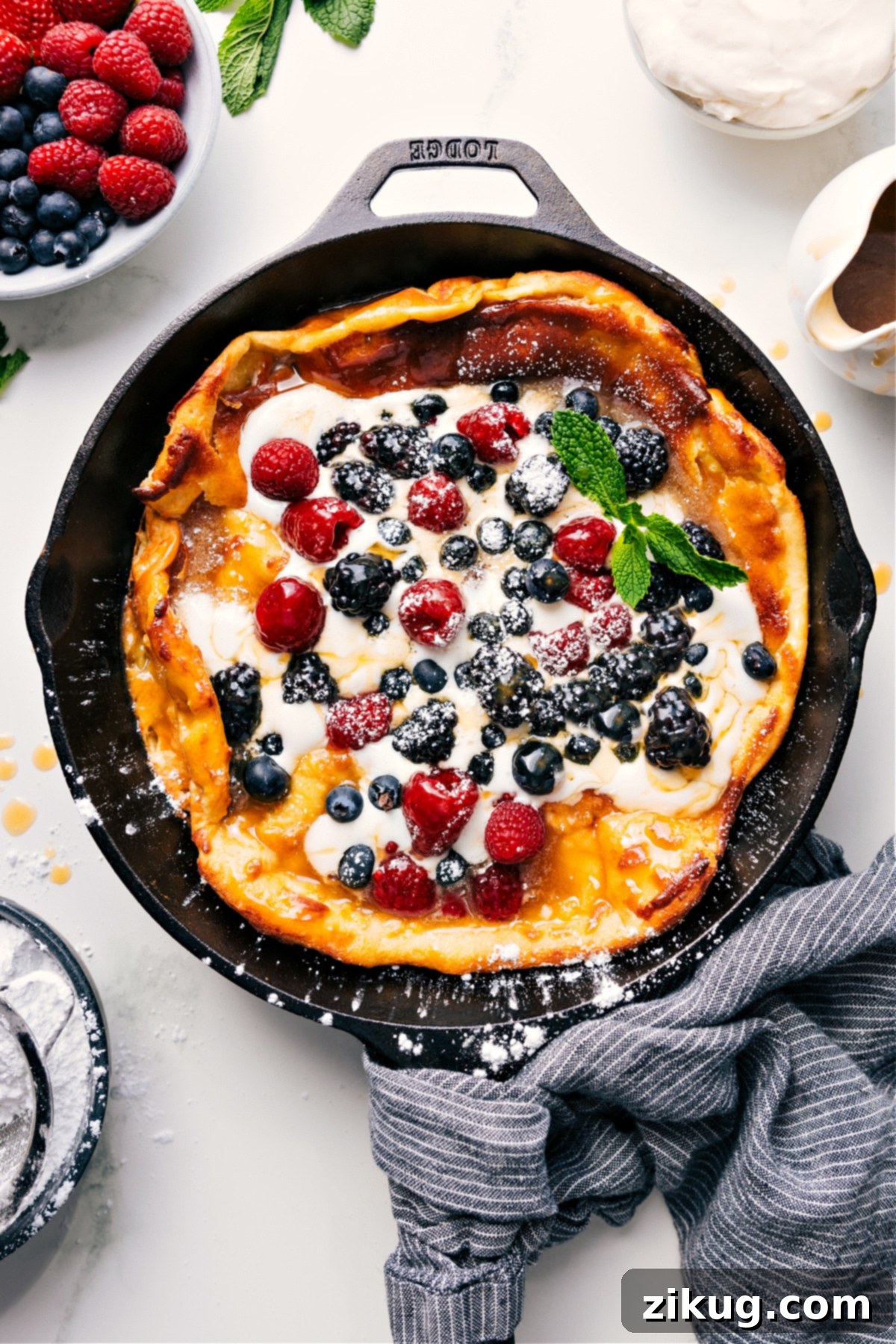 The Best Dutch Baby Recipe Dutch Baby skillet topped with fresh whipped cream, an assortment of berries, and a generous drizzle of syrup, ready to be served.