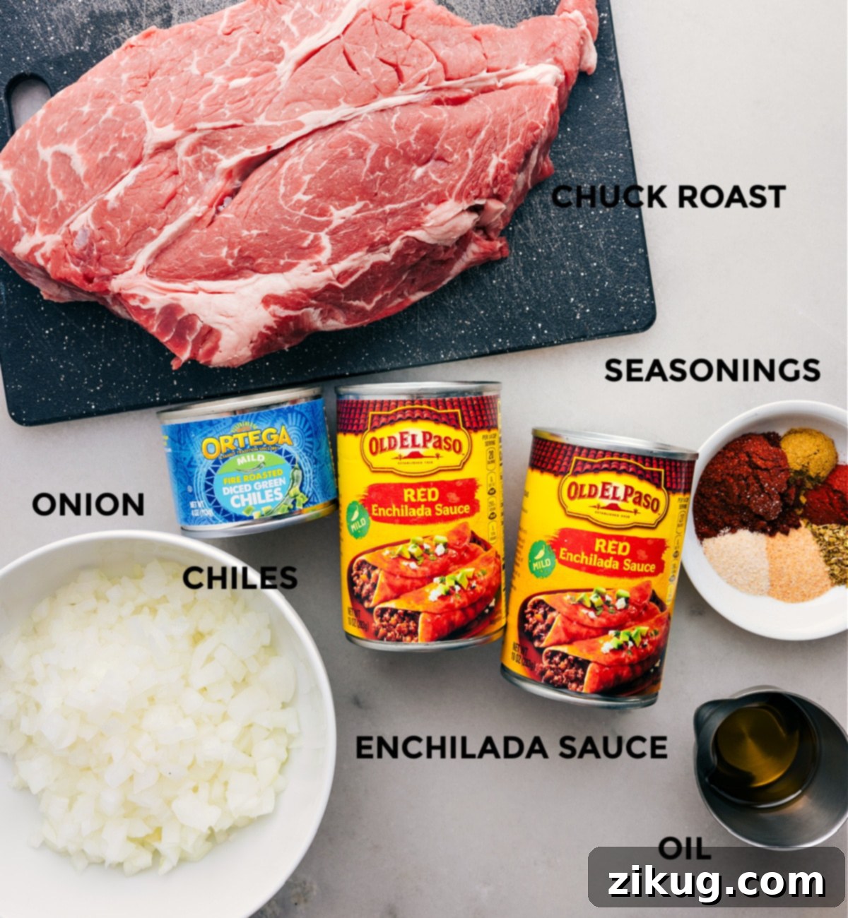 All the essential ingredients for shredded beef tacos laid out: beef chuck roast, olive oil, yellow onion, various taco seasonings, mild red enchilada sauce, and diced green chiles.