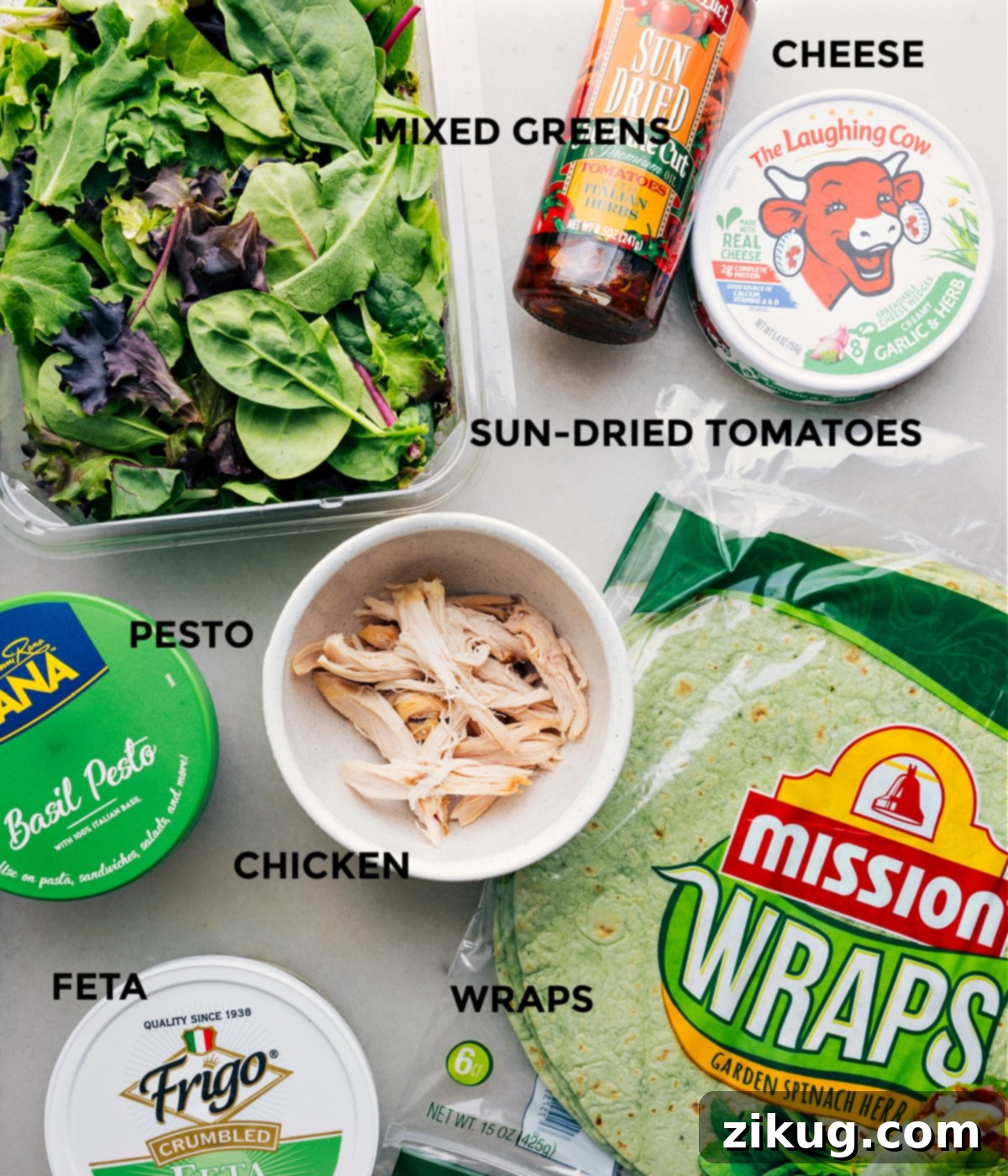 A beautiful display of all the fresh ingredients for a Mediterranean Wrap laid out for easy assembly: a tortilla, vibrant pesto, shredded rotisserie chicken, mixed greens, sun-dried tomatoes, Laughing Cow cheese, and crumbled feta.