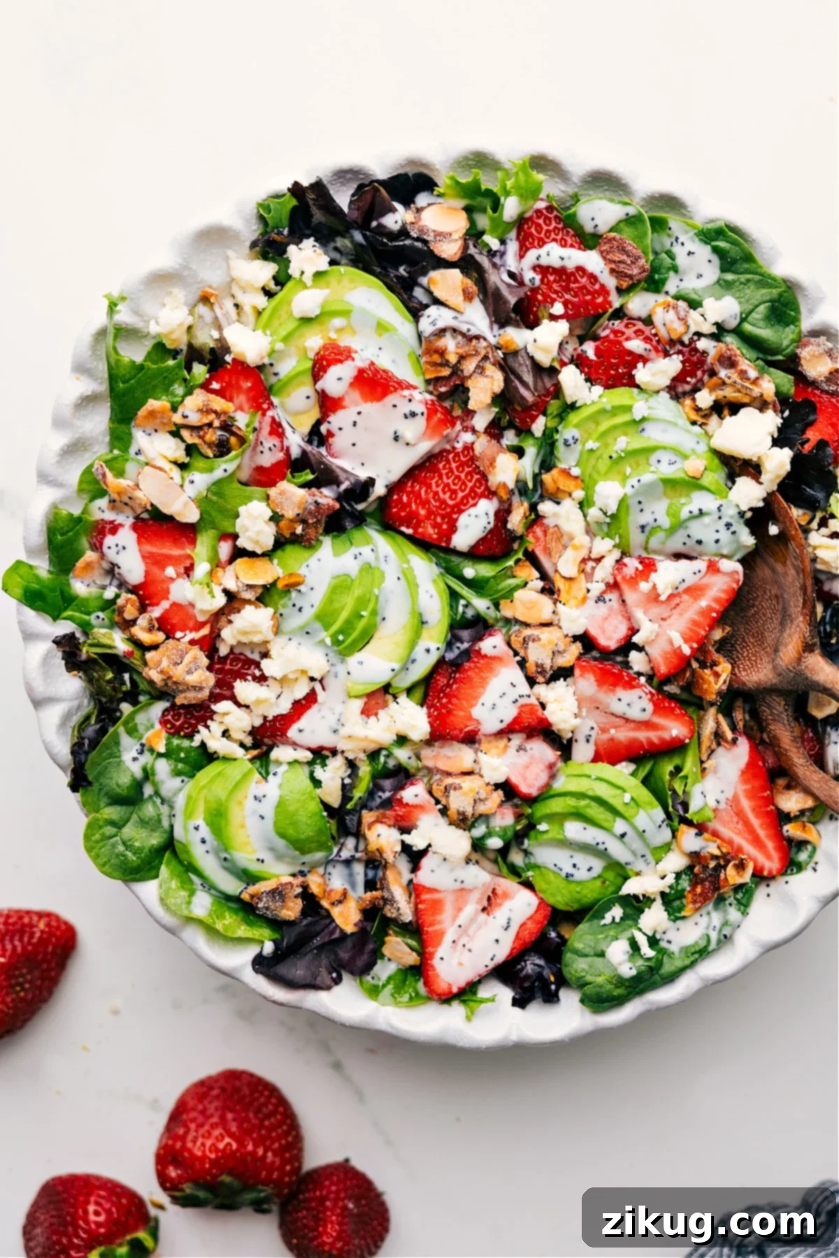 Colorful and Fresh Strawberry Poppy Seed Salad A vibrant Strawberry Poppyseed Salad tossed in a large white bowl, garnished with fresh mint, drizzled with a creamy poppyseed dressing, and ready to be served. The salad features sliced red strawberries, diced green avocado, crumbled white feta cheese, and golden candied almonds, all resting on a bed of mixed greens.
