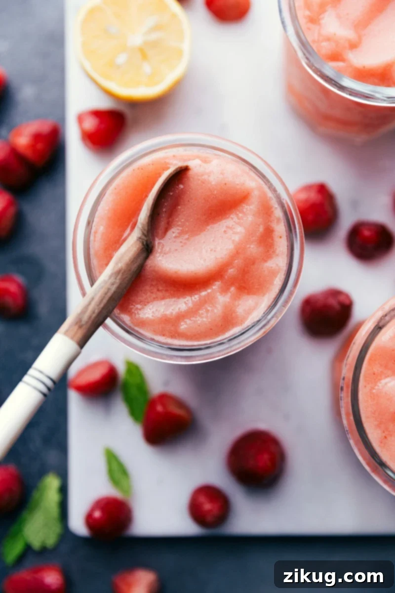 Frosty and refreshing Lemon Berry Slush with a spoon, perfect for a hot day. Refreshing homemade lemon berry slush in a clear glass with a spoon, garnished with a fresh strawberry and a lemon slice, ready to be enjoyed.