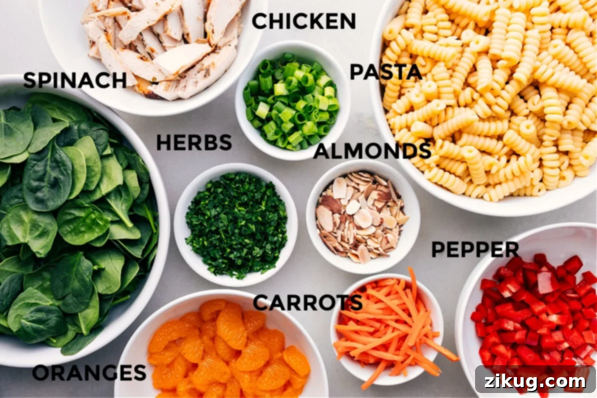 Asian pasta salad ingredients laid out for easy assembly including chicken, pasta, almonds, herbs, spinach, peppers, carrots, and oranges.