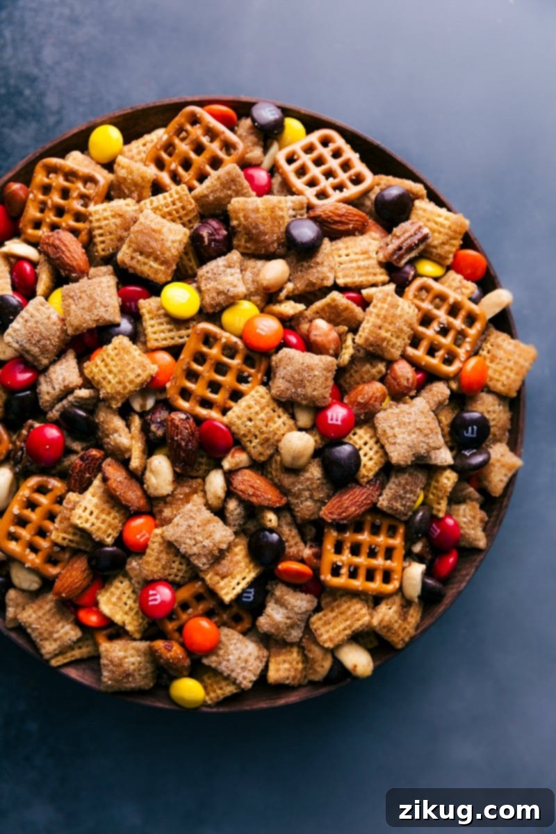 Crunchy Pumpkin Spice Snack Mix with cinnamon cereal, mixed nuts, pretzels, and M&M's, perfect for fall and holidays. Overhead view of a generous bowl of Pumpkin Spice Snack Mix, showing cinnamon cereals, mixed nuts, pretzels, and autumn-colored candies coated in a golden caramel glaze.