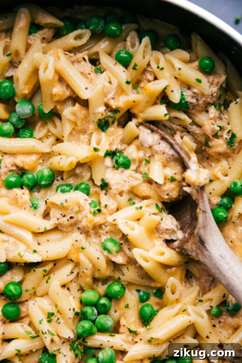 Finished Tuna Pasta in Skillet - Spoon Scooping a Serving A finished skillet of cheesy tuna pasta with peas, a spoon poised to serve a hearty portion, highlighting the dish's readiness.