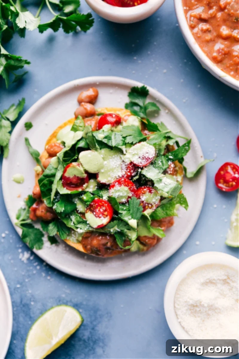 Deliciously Crispy Bean Tostadas: A Simple & Flavorful Recipe with Smashed Avocado, Spiced Pinto Beans, Fresh Veggies, and a Zesty Cilantro-Lime Sauce. A Chelsea's Messy Apron Creation. Overhead shot of crispy Bean Tostadas, generously topped with smashed avocado, seasoned pinto beans, fresh lettuce, juicy tomatoes, and cotija cheese, finished with a cilantro-lime sauce.
