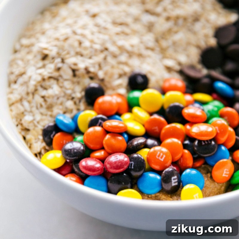 Flourless Monster Cookies made with a few healthier swaps Oats, chocolate chips, and M&M's being added to the bowl of wet ingredients, showcasing the final stage of dough preparation for flourless monster cookies.