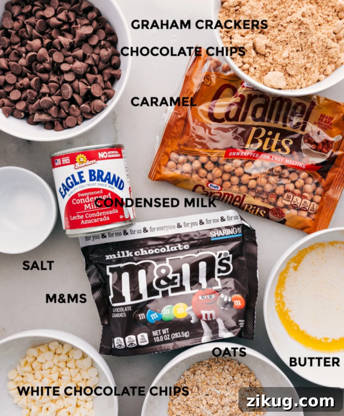 All the ingredients are prepped and ready for easy assembly: graham crackers, caramel bits, condensed milk, chocolate chips, M&Ms, butter, oats, and salt, neatly arranged.