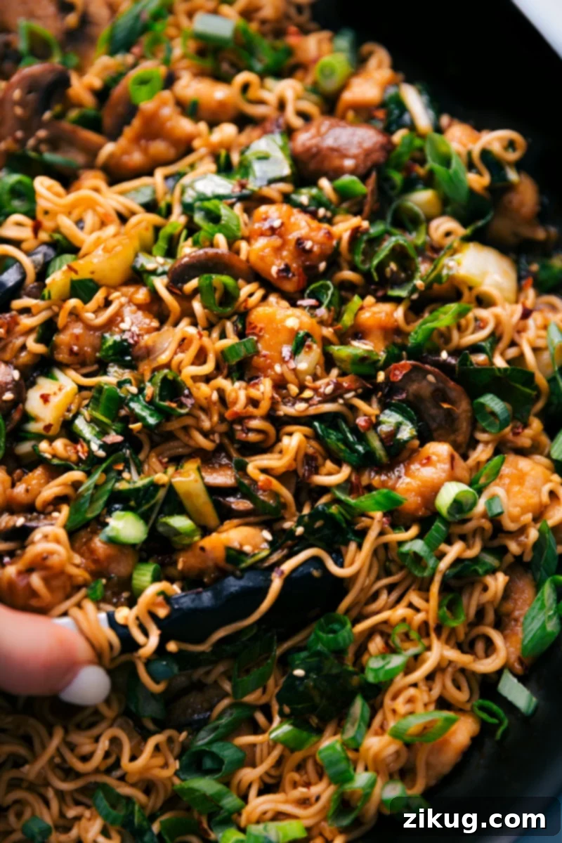 A large pot filled with General Tso Ramen, showing noodles, chicken, and vegetables, with tongs lifting a serving.