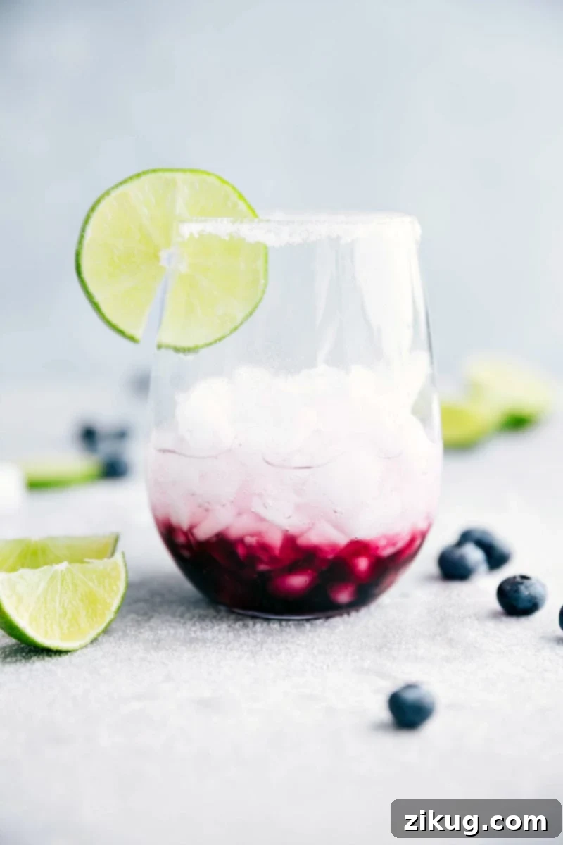 Chilled Limeberry Fizz 3 Close-up of a glass of blueberry mandarin-lime spritzer with ice, fresh blueberries, mint, and a sugar-rimmed glass, highlighting its refreshing appeal.